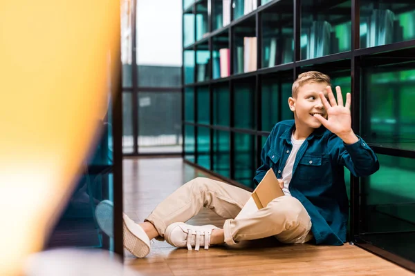 Smiling schoolboy holding book and waving hand while sitting on floor in library — Stock Photo