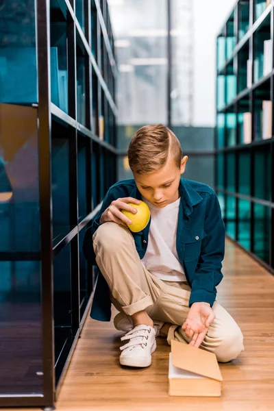 Adorable colegial sosteniendo manzana y leer libro mientras está sentado en el suelo en la biblioteca - foto de stock