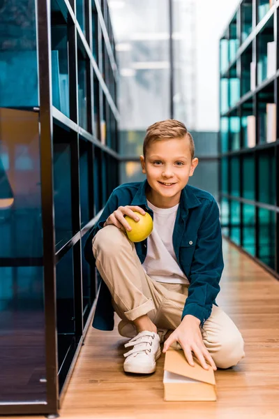 Adorabile scolaro che tiene mela e sorride alla macchina fotografica mentre siede sul pavimento in biblioteca — Foto stock