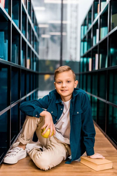 Lindo colegial sosteniendo manzana y sonriendo a la cámara mientras está sentado en el suelo en la biblioteca - foto de stock