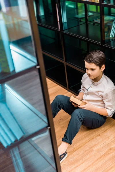 Blick von oben auf den nachdenklichen Jungen, der Buch in der Hand hält und in der Bibliothek auf dem Boden sitzt — Stockfoto