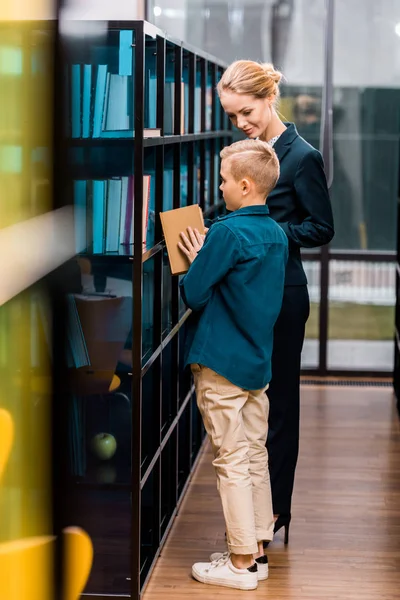 Lächelnde junge Bibliothekarin schaut den Jungen an, der Buch in der Bibliothek hält — Stockfoto