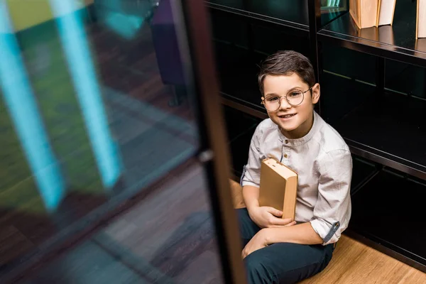 Visão de alto ângulo de estudante sorrindo em óculos segurando livro e sentado no chão na biblioteca — Fotografia de Stock