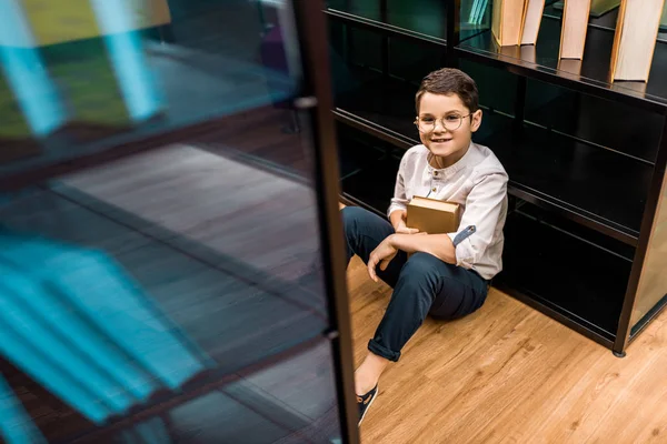 High angle view of schoolboy in eyeglasses holding book and smiling at camera in library — Stock Photo