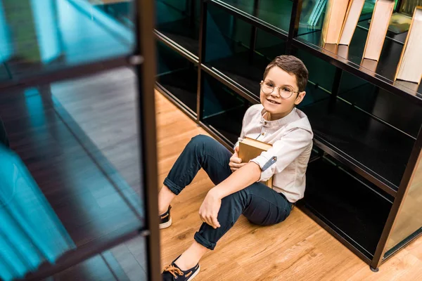 Vista ad alto angolo del ragazzo con gli occhiali che tiene il libro e sorride alla fotocamera in biblioteca — Foto stock