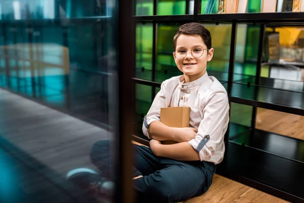 Cute smiling schoolboy in eyeglasses holding book and sitting on floor in library — Stock Photo