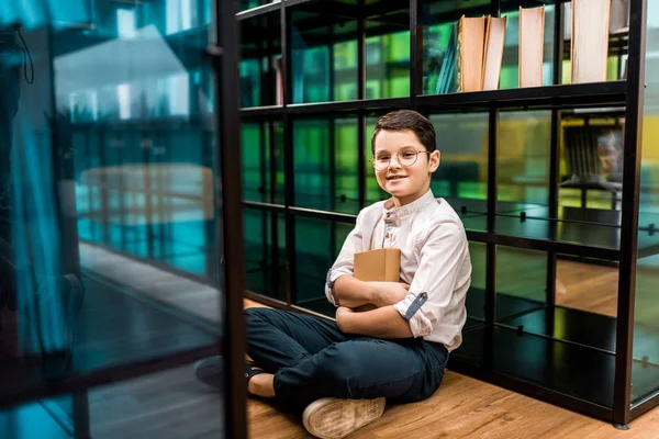 Cute happy schoolboy in eyeglasses holding book and sitting on floor in library — Stock Photo