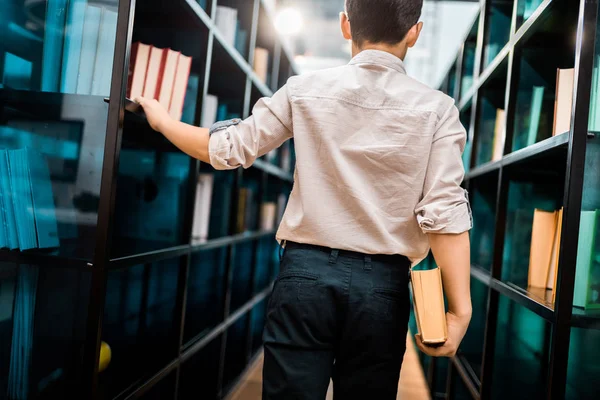 Back view of boy holding book and walking between bookshelves in library — Stock Photo