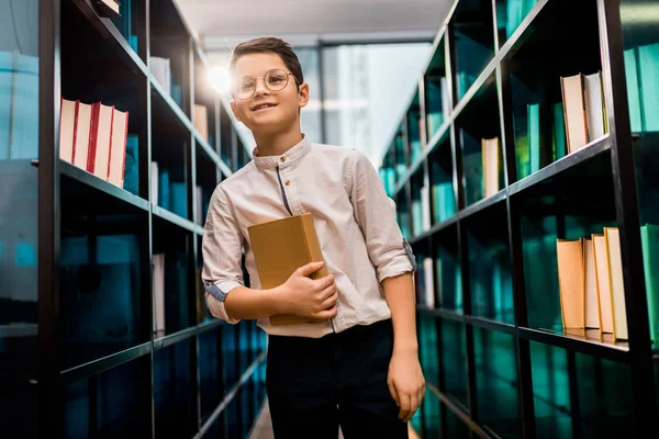 Adorabile sorridente scolaro in occhiali che tiene libro in biblioteca — Foto stock