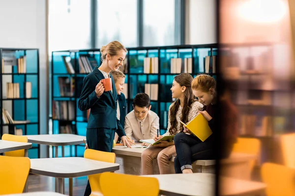 Souriant jeune bibliothécaire tenant tasse et regardant les écoliers à la bibliothèque — Photo de stock