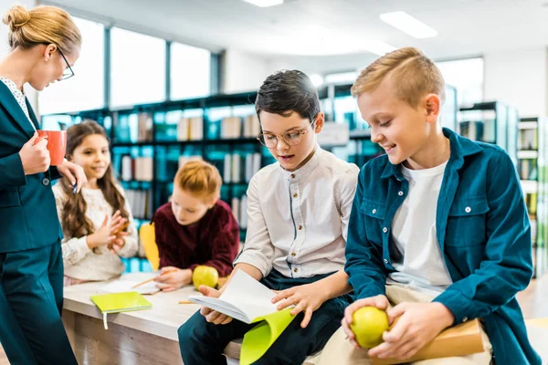 Young female librarian looking at kids reading books in library — Stock Photo