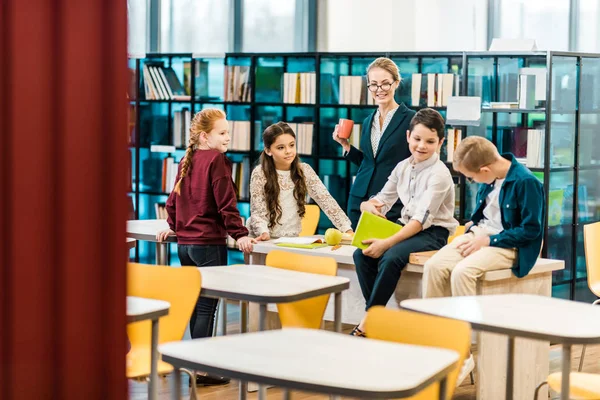 Adoráveis crianças em idade escolar e jovem bibliotecária em pé e sentado na biblioteca — Fotografia de Stock