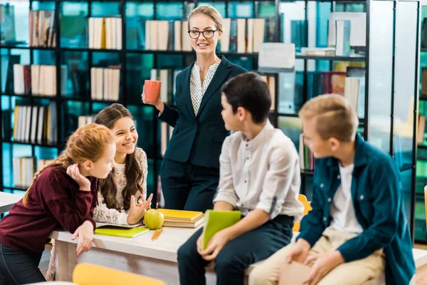 Junge Bibliothekarin hält Becher in der Hand und schaut lächelnde Schulkinder in der Bibliothek an — Stockfoto