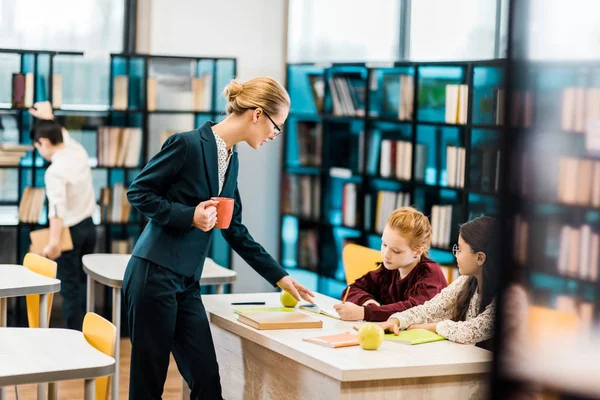 Giovane insegnante femminile in possesso di tazza e guardando le studentesse che studiano in biblioteca — Foto stock