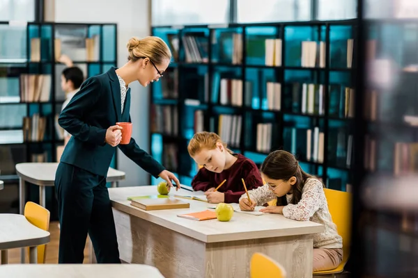 Jeune enseignante tenant une tasse et regardant des écoliers étudier à la bibliothèque — Photo de stock
