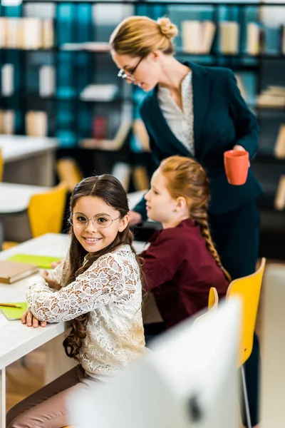 Чарівний школярка посміхається на камеру під час навчання з однокласником і вчителем в бібліотеці — стокове фото