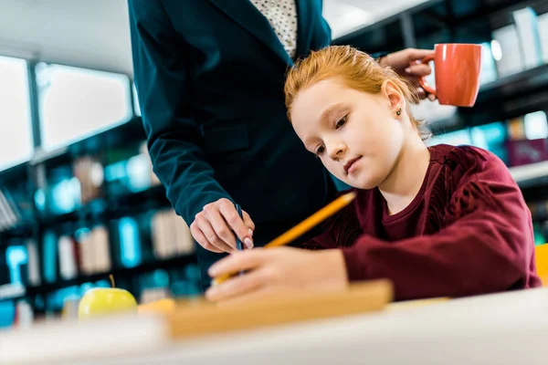 Cropped shot of teacher holding cup and helping schoolgirl studying in library — Stock Photo