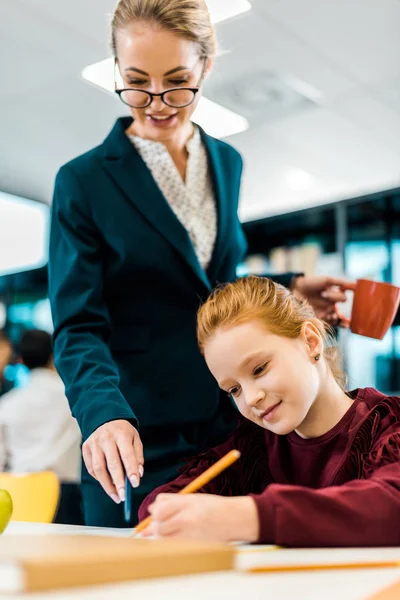 Smiling young teacher in eyeglasses looking at schoolgirl studying in library — Stock Photo