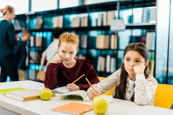 Schoolkids writing with pencils and looking away in library — Stock Photo
