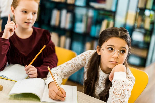 Adorable schoolgirls writing with pencils and looking away while studying in library — Stock Photo