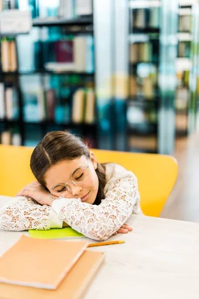 Lindo escolar en gafas durmiendo en la mesa con libros en la biblioteca - foto de stock