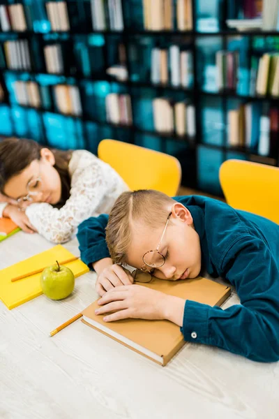 Cute tired schoolkids in eyeglasses sleeping on table with books in library — Stock Photo