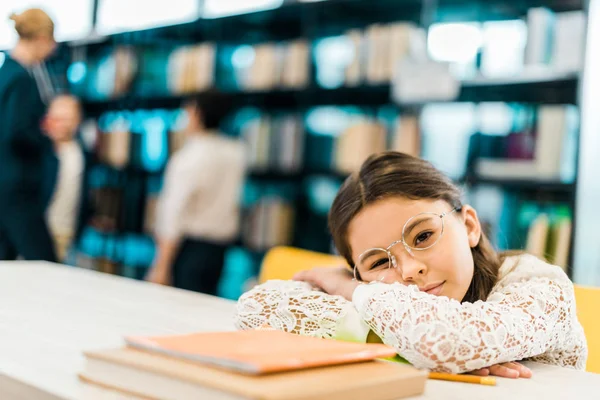 Скучающая школьница в очках, прислонившаяся к столу с книгами и смотрящая на камеру в библиотеке — стоковое фото