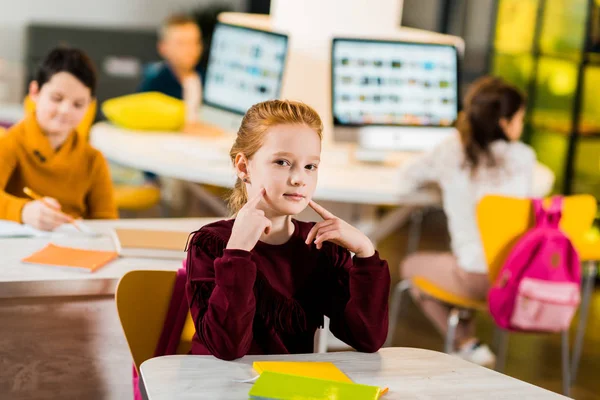 Entzückendes Schulkind blickt in die Kamera, während es mit Klassenkameraden in der Bibliothek sitzt — Stockfoto