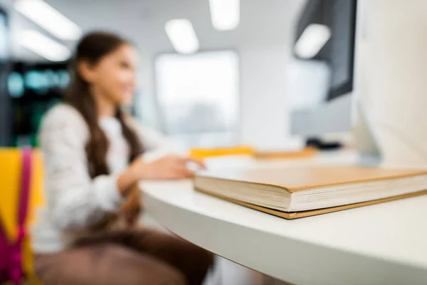 Vue rapprochée du livre et de l'écolière en utilisant l'ordinateur de bureau derrière dans la bibliothèque — Photo de stock