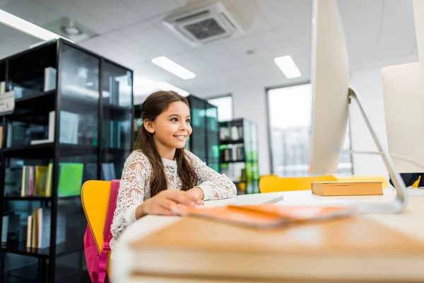 Adorable smiling schoolgirl using desktop computer in library — Stock Photo