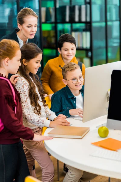 Cute schoolchildren and librarian using computer together in library — Stock Photo