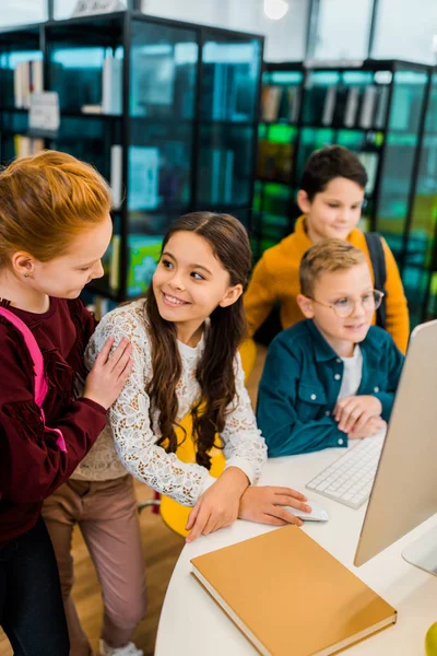 Schöne glückliche Schulkinder, die gemeinsam einen Desktop-Computer in der Bibliothek benutzen — Stockfoto