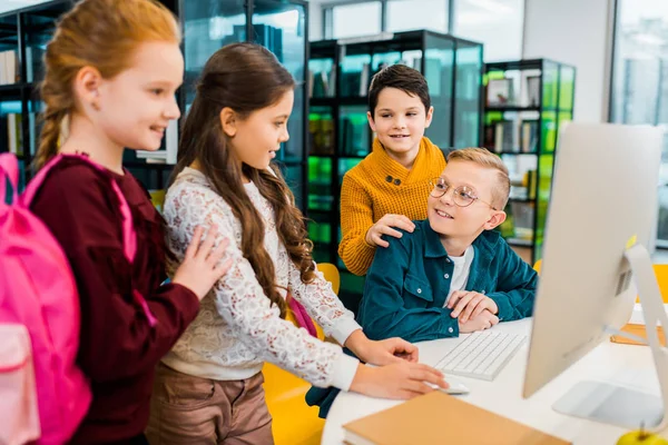 Cute smiling schoolchildren using desktop computer together in library — Stock Photo