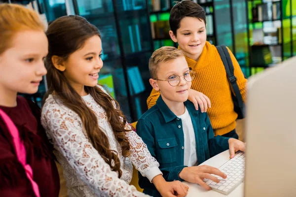 Cute happy schoolkids using desktop computer in library — Stock Photo