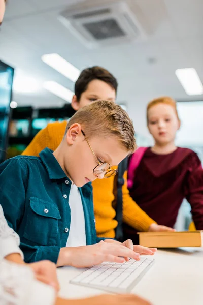 Crianças olhando menino digitando no teclado do computador na biblioteca — Fotografia de Stock