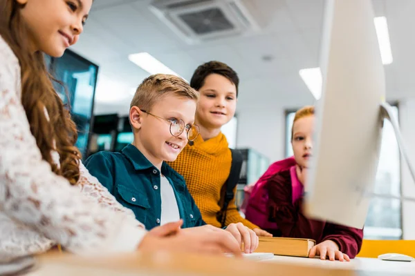Очаровательные улыбающиеся школьники, использующие настольный компьютер в библиотеке — стоковое фото