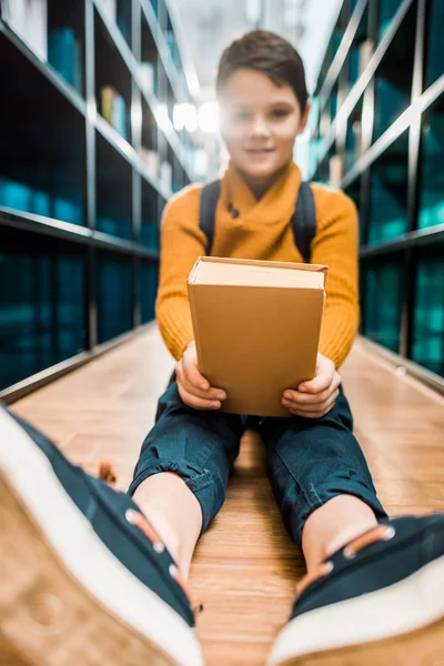 Estudante segurando livro e sorrindo para a câmera enquanto sentado no chão na biblioteca — Fotografia de Stock