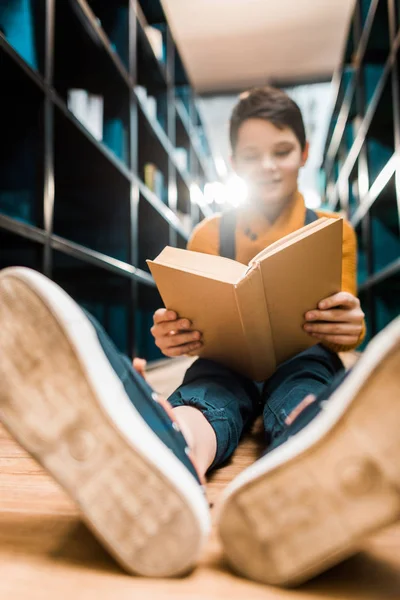 Selective focus of schoolboy reading book and sitting on floor in library — Stock Photo