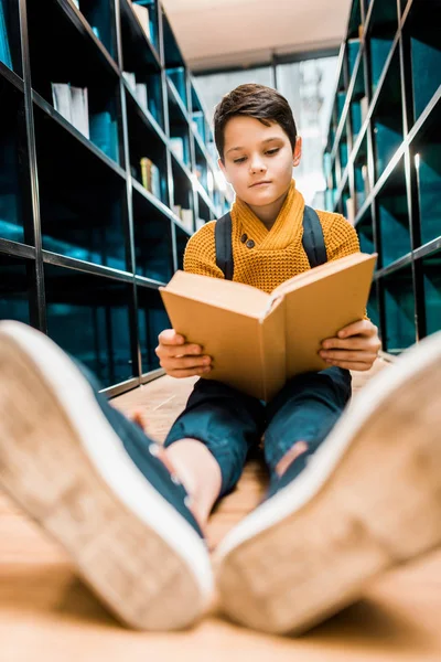 Colegial sentado en el suelo y leyendo libro en la biblioteca - foto de stock