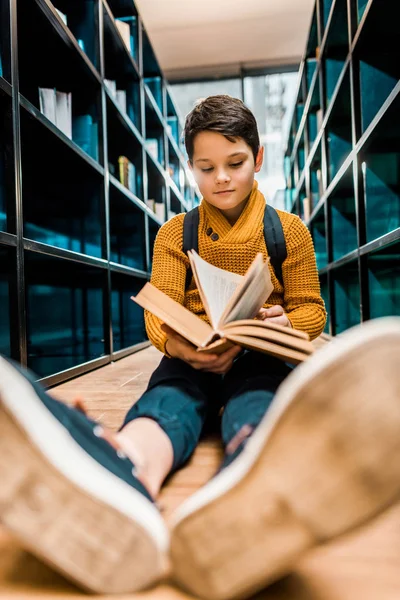 Entzückend lächelnder Schuljunge liest Buch und sitzt auf dem Fußboden in der Bibliothek — Stockfoto