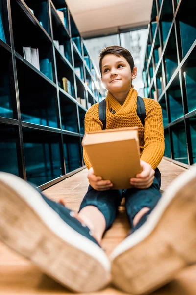 Adorable smiling schoolboy holding book and sitting on floor in library — Stock Photo