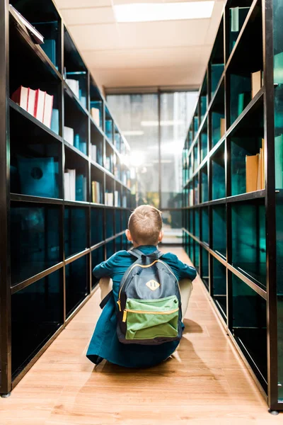 Вид сзади школьника с рюкзаком, сидящего на полу в библиотеке — стоковое фото