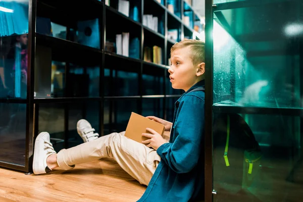 Вид збоку мила школярка тримає книгу і дивиться геть, сидячи на підлозі в бібліотеці — стокове фото