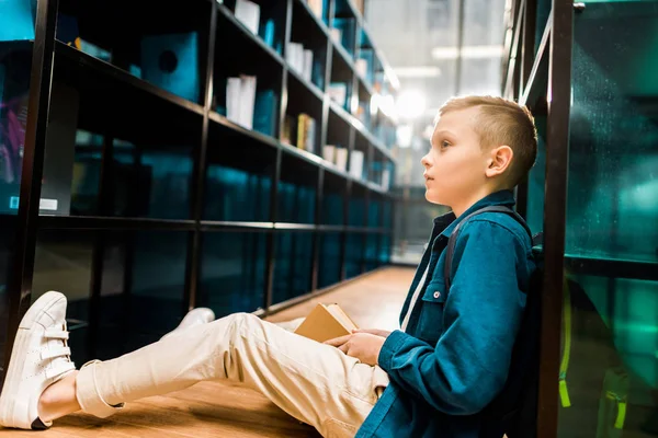 Side view of cute schoolboy holding book and looking away while sitting on floor in library — Stock Photo