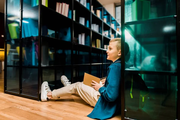 Vista lateral do menino sorridente sentado no chão e livro de leitura na biblioteca — Fotografia de Stock