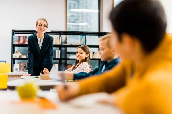 Smiling young teacher looking at schoolchildren studying in library — Stock Photo