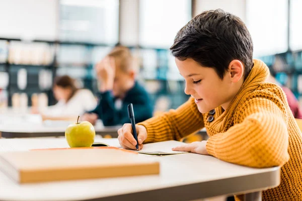 Усміхнений хлопчик пише з ручкою під час навчання з однокласниками в бібліотеці — стокове фото