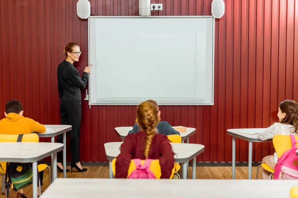 Profesor sonriente apuntando a pizarra interactiva y mirando a los escolares - foto de stock