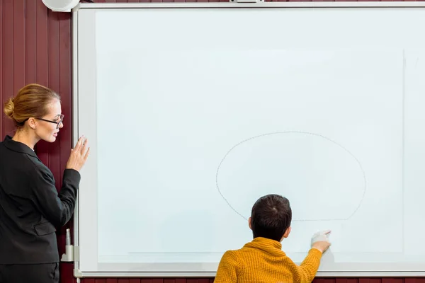 Joven profesor mirando a colegial estudiando con pizarra interactiva - foto de stock