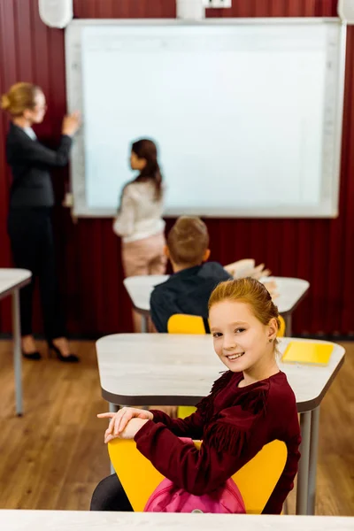 Vista de ángulo alto de adorable escolar sonriendo a la cámara mientras sus compañeros de clase y profesor estudian con pizarra blanca detrás - foto de stock